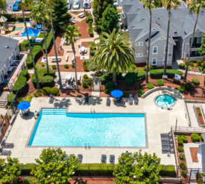 aerial view of large swimming pool and hot tub, gated, multiple picnic tables, picnic area outside pool area as well, handicap accessible ramp to pool area, large palm trees surrounding pool area, photo taken on a sunny day