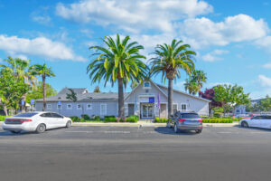 Exterior Leasing Office, beautiful palm trees in front of entrance, meticulously groomed grounds, parking out front, photo taken on a sunny day.