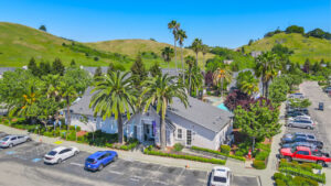 Aerial View of Leasing Office, palm trees in front of entrance, parking out front, trees scattered throughout property, sprawling hills in the distance.